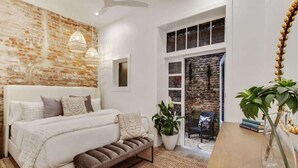 A bedroom with a brick wall, a white bed, a bench, a potted plant, and a wooden table with a vase.
