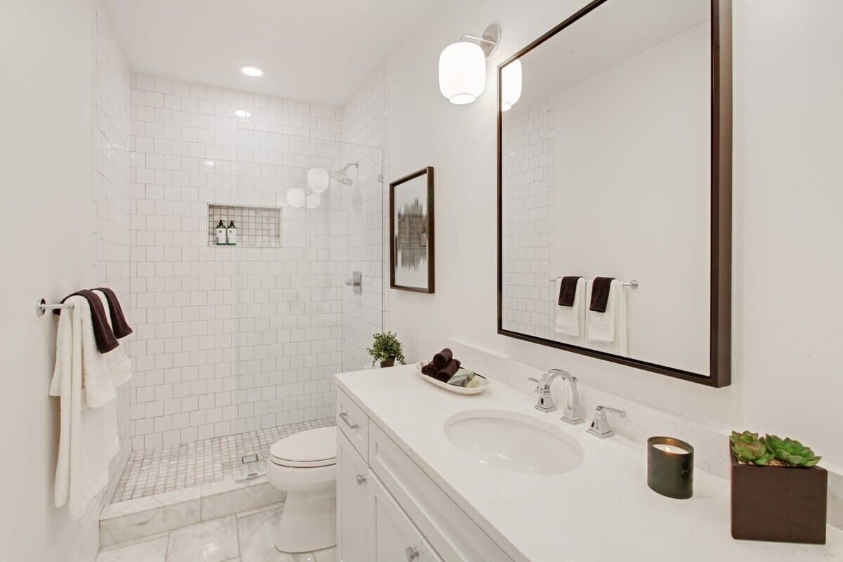 Un baño moderno con azulejos blancos, un espejo grande, un lavamanos y un inodoro.