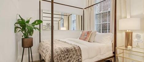 A four-poster canopy bed with a white duvet, a patterned throw, and a small potted plant beside it.