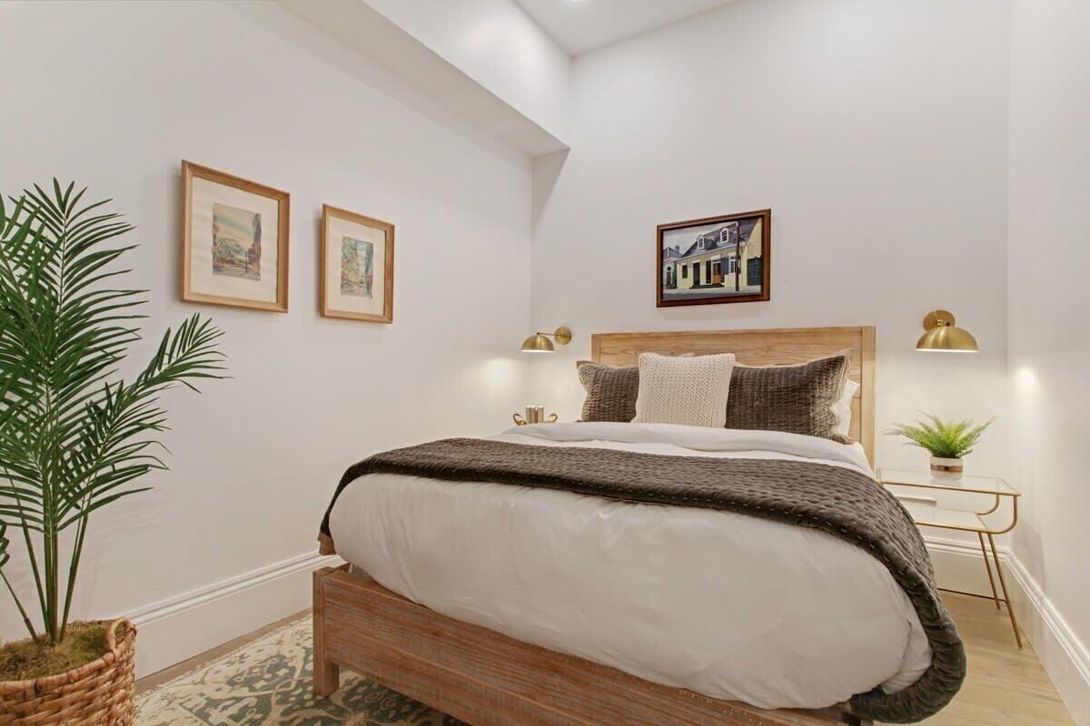 Une chambre avec un lit en bois, une grande plante en pot, des tableaux encadrés accrochés au mur et une petite table de chevet avec une lampe.