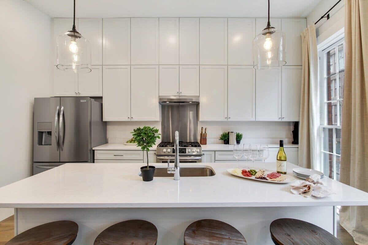 Una cocina moderna con armarios blancos, electrodomésticos de acero inoxidable, una isla central con taburetes, iluminación colgante y un amplio ventanal con cortinas.