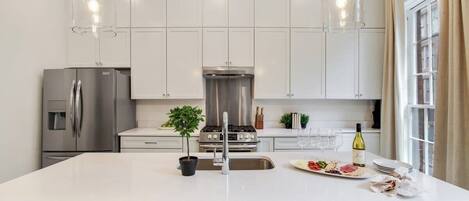 Una cocina moderna con armarios blancos, electrodomésticos de acero inoxidable, una isla central con taburetes, iluminación colgante y un amplio ventanal con cortinas.