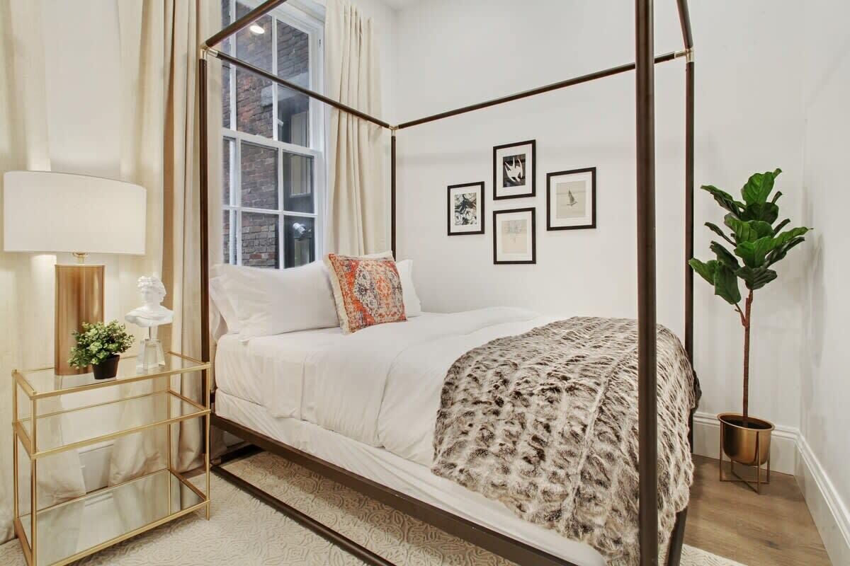 A four-poster bed with a white and patterned bedspread, a bedside table with a lamp, and a potted plant.