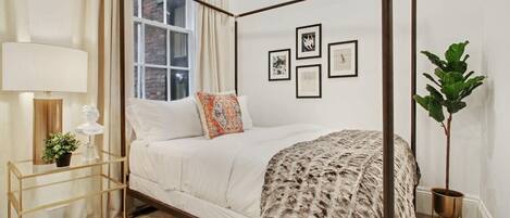 A four-poster bed with a white and patterned bedspread, a bedside table with a lamp, and a potted plant.