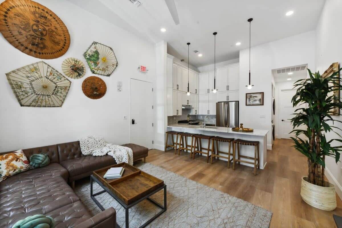 A modern living room with a brown leather sofa, a wooden coffee table, and a kitchen area with white cabinets and wooden stools.