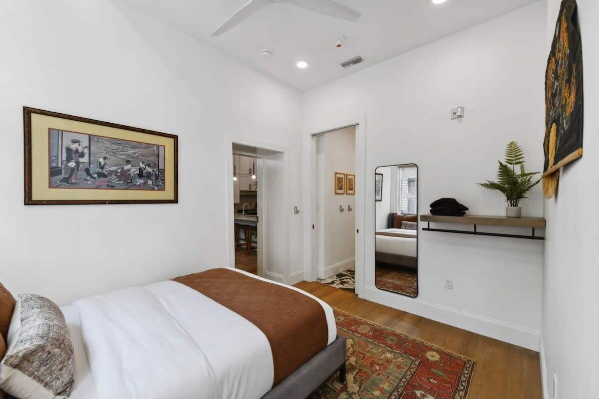 A bedroom with a bed, a bedside table, a mirror, a framed picture on the wall, and a door leading to another room.
