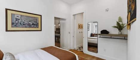 A bedroom with a bed, a bedside table, a mirror, a framed picture on the wall, and a door leading to another room.
