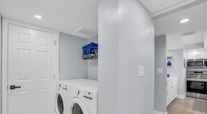 A laundry room with a washing machine and dryer, a shelf with folded clothes, and a kitchen area with a microwave and oven. - E-234 & F- 237 - Perfect for Group Travel (Naples)