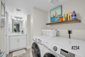 A modern laundry room with a Samsung washer and dryer, a mirror, a sink, and a shelf with cleaning supplies.
