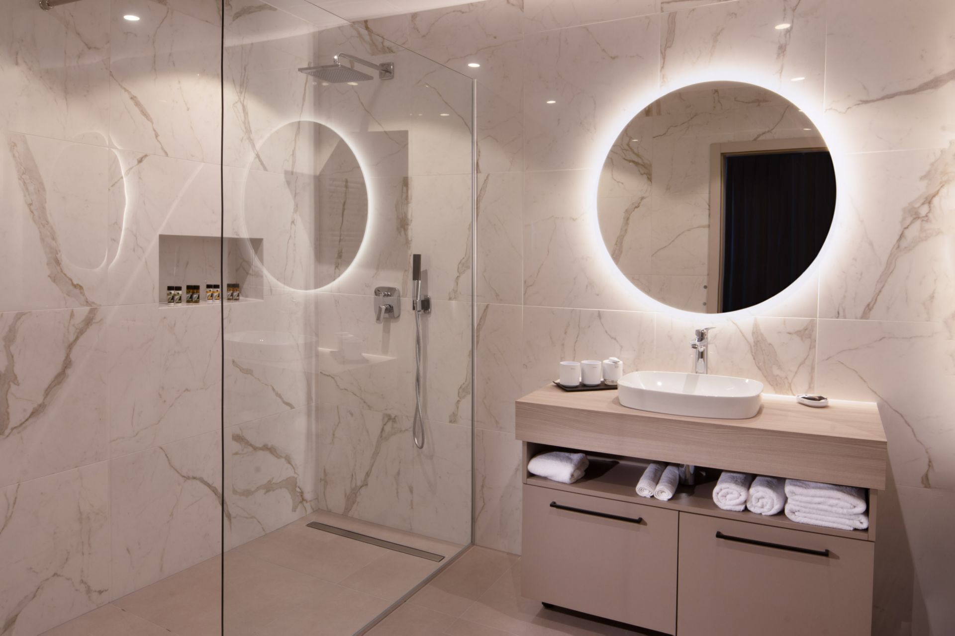 Een moderne badkamer met een douchecabine van glas, een ronde spiegel en een wastafelmeubel met een wastafel en opbergruimte voor handdoeken.