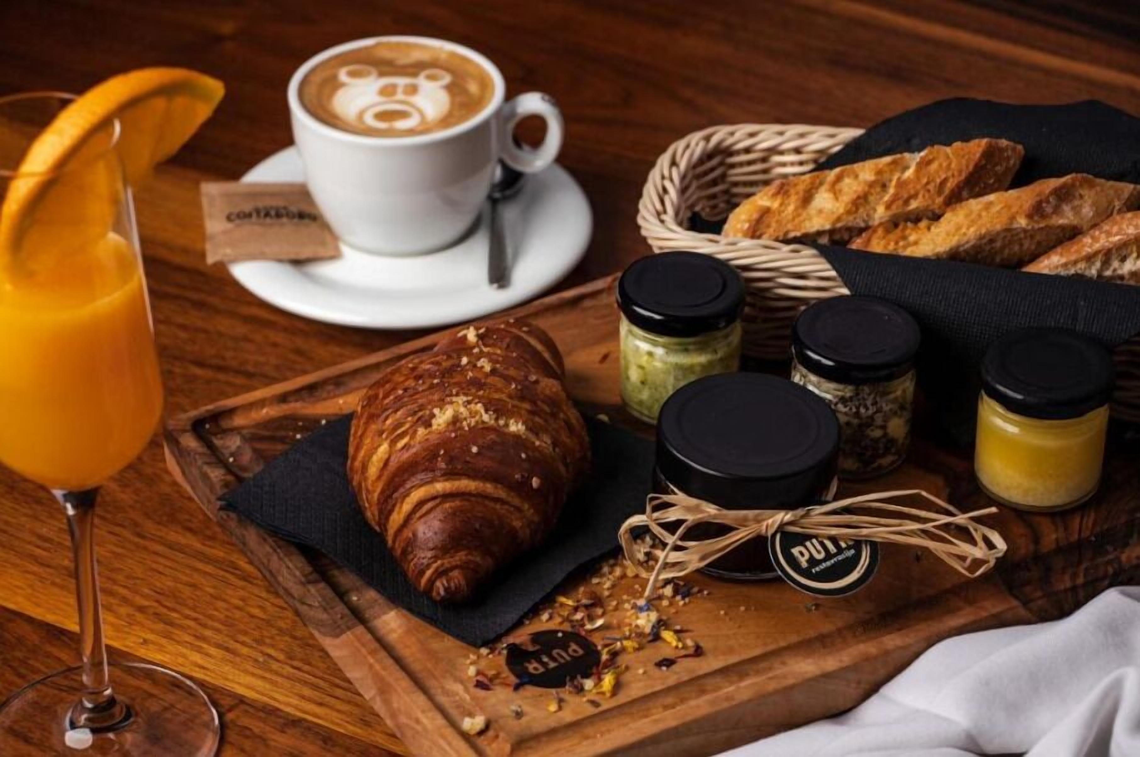 Ein Frühstücksbuffet mit einem Croissant, Kaffee, Orangensaft und verschiedenen Beilagen auf einem Holbrett.