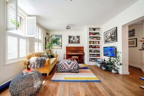 Un salon avec un canapé, un pouf et un piano. On y trouve également des étagères et un téléviseur.