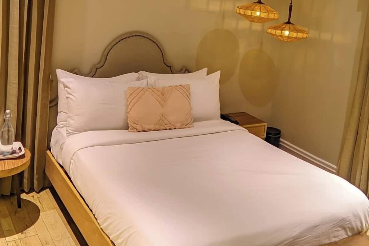 A neatly made bed with white linens and a wooden headboard, a small bedside table with a bottle, and a bench at the foot of the bed.