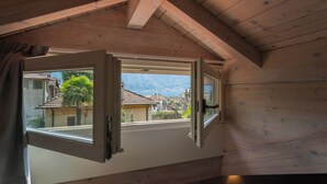 A room with a wooden ceiling and a window with an open shutter, revealing a view of houses and mountains. - Villa Olga With Private Pool by Rent all Como (Tremezzina)