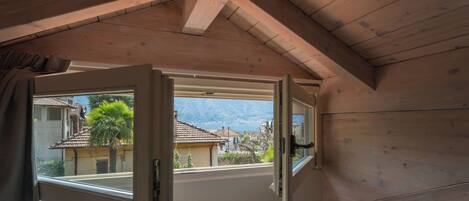 Una habitación con techo de madera y una ventana con contraventana abierta, mostrando vistas de casas y montañas.