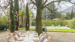 An outdoor dining area with a table set for six, surrounded by chairs, in a garden with a pool and lounge chairs in the background. - Villa Olga With Private Pool by Rent all Como (Tremezzina)