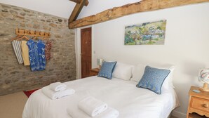 Un dormitorio con pared de piedra, vigas de madera, cama con ropa de cama blanca y almohadas azules, un cuadro y una mesita de noche de madera.
