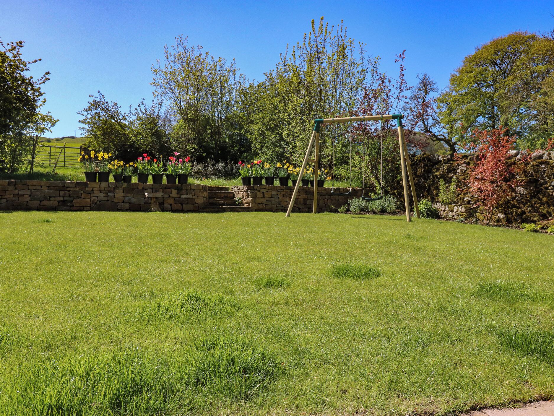 A grassy backyard with a stone wall, a swing set, and flowering plants.