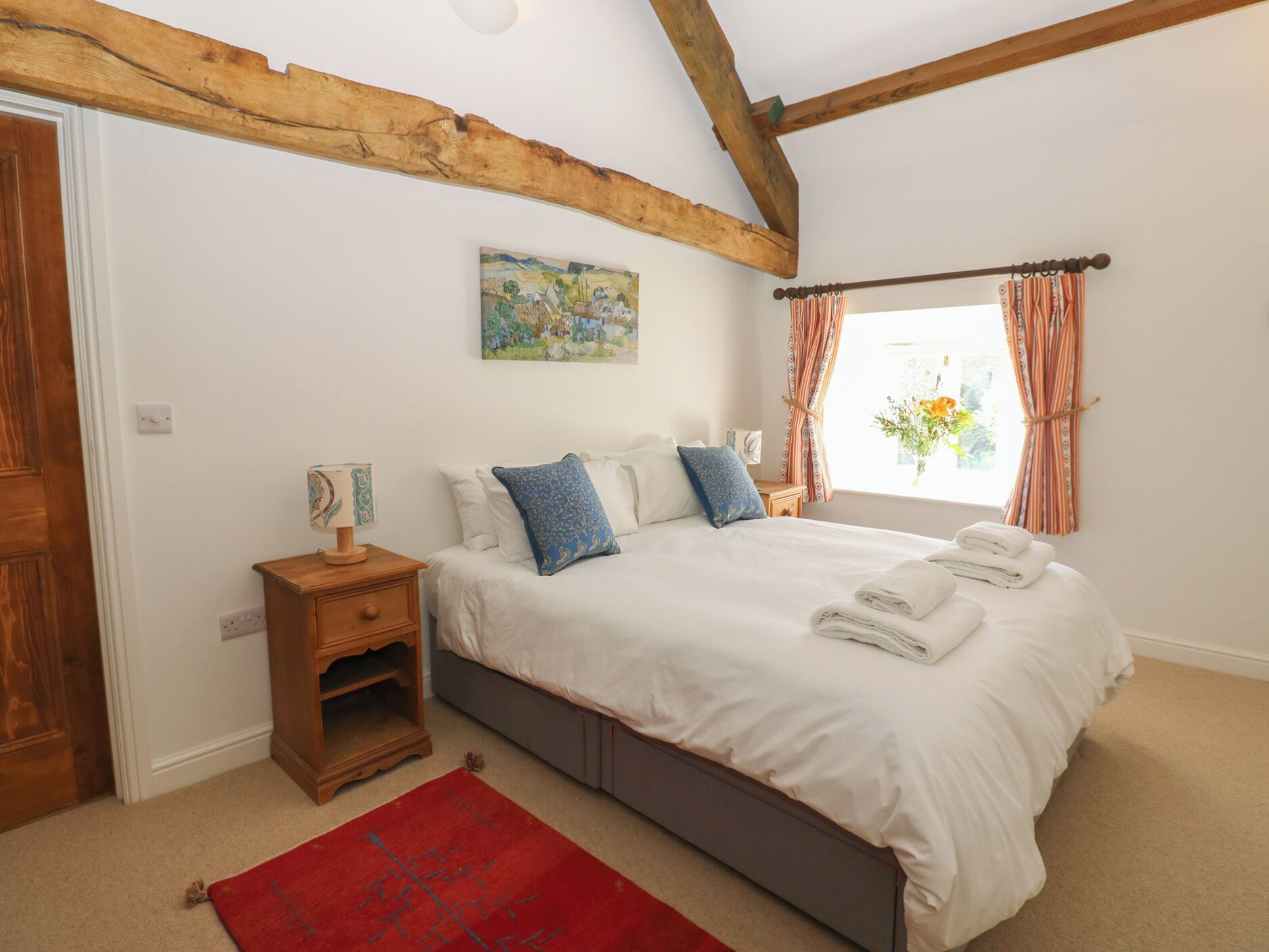 A bedroom with a bed, a nightstand, a red rug, a painting, and a window with curtains.