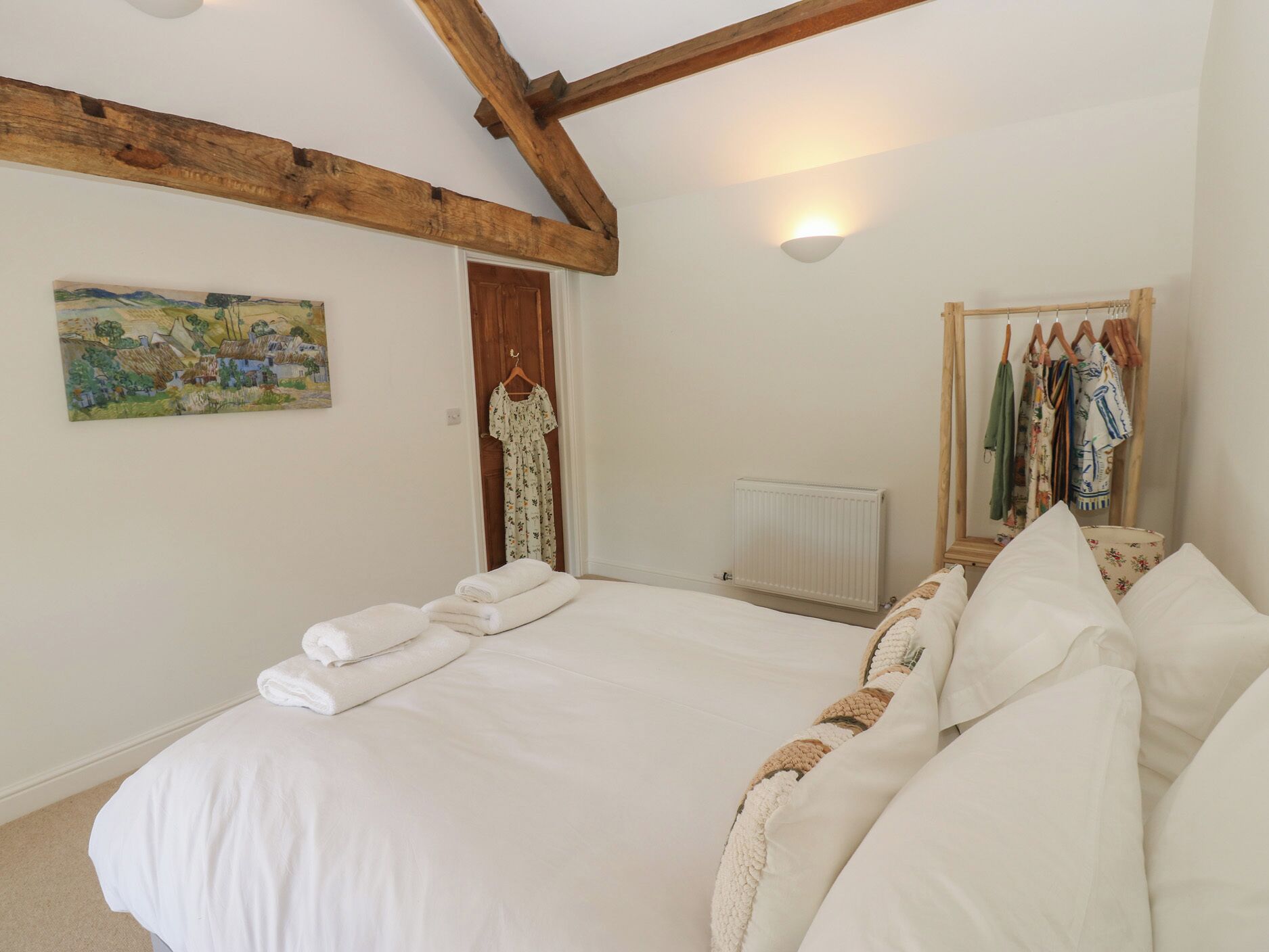 A bedroom with a large bed, a radiator, a wooden ceiling beam, a painting, and a clothing rack.