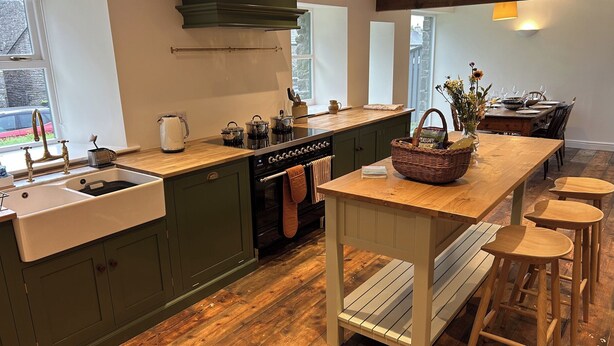Una cocina moderna con suelo de madera, armarios verdes, una isla central con taburetes y una zona de comedor con una mesa preparada para cuatro.