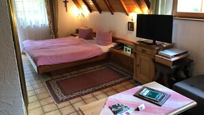 Un dormitorio con cama, mesita de noche, televisor y escritorio con computadora y libros.