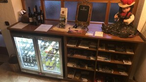 Una zona de bar con un refrigerador lleno de bebidas, un expositor con libros y un adorno que parece una muñeca.