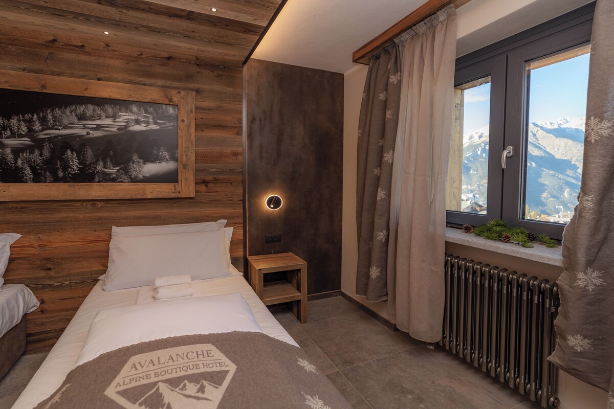 Una habitación de hotel con techo de madera, una cama con manta de motivos montañosos, una luz nocturna, una ventana con vistas a las montañas y un radiador.