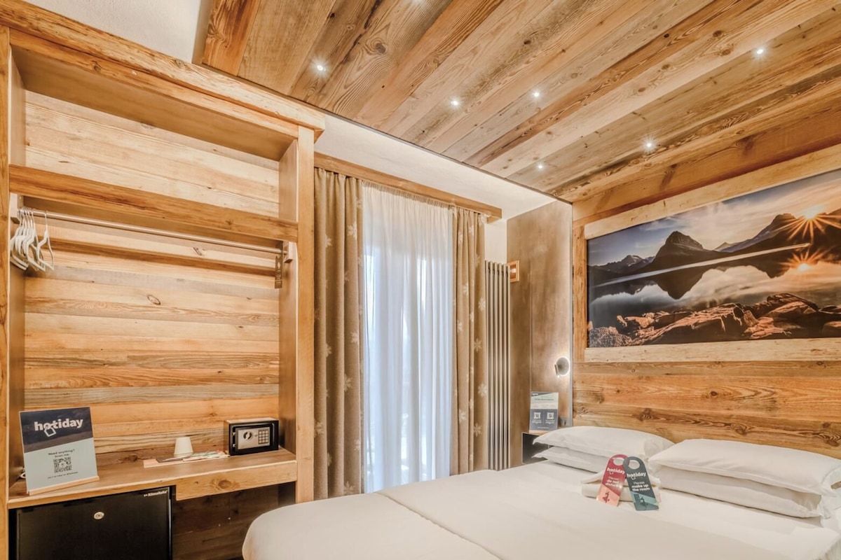 Habitación de hotel con techo de madera, cama con ropa de cama blanca, pintura de un paisaje montañoso y escritorio con computadora y un cartel que dice "hotday".
