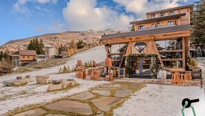 Una cabaña de madera con un patio de piedra, rodeada de nieve y montañas.