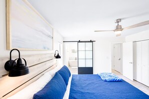 A bedroom with a bed, a ceiling fan, a nightstand, a wall-mounted lamp, and a sliding door leading to a bathroom. - Butler Beach 3BR | Fireplace + Private Backyard! (St. Augustine)