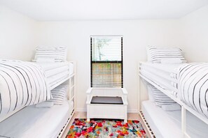 A bedroom with two single beds, a window with blinds, a colorful rug, and a small white bench.