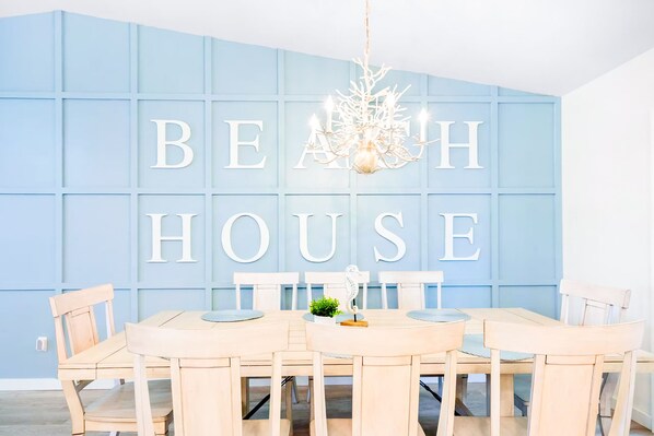 A dining area with a table and chairs, a chandelier, and a wall with the words 'BEACH HOUSE' in large letters.