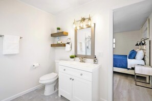 A modern bathroom with a white toilet, vanity with drawers, a mirror with lights, and a shower area visible through an open door.
