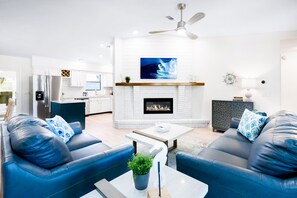 A modern living room with a fireplace, a ceiling fan, and a kitchen in the background.