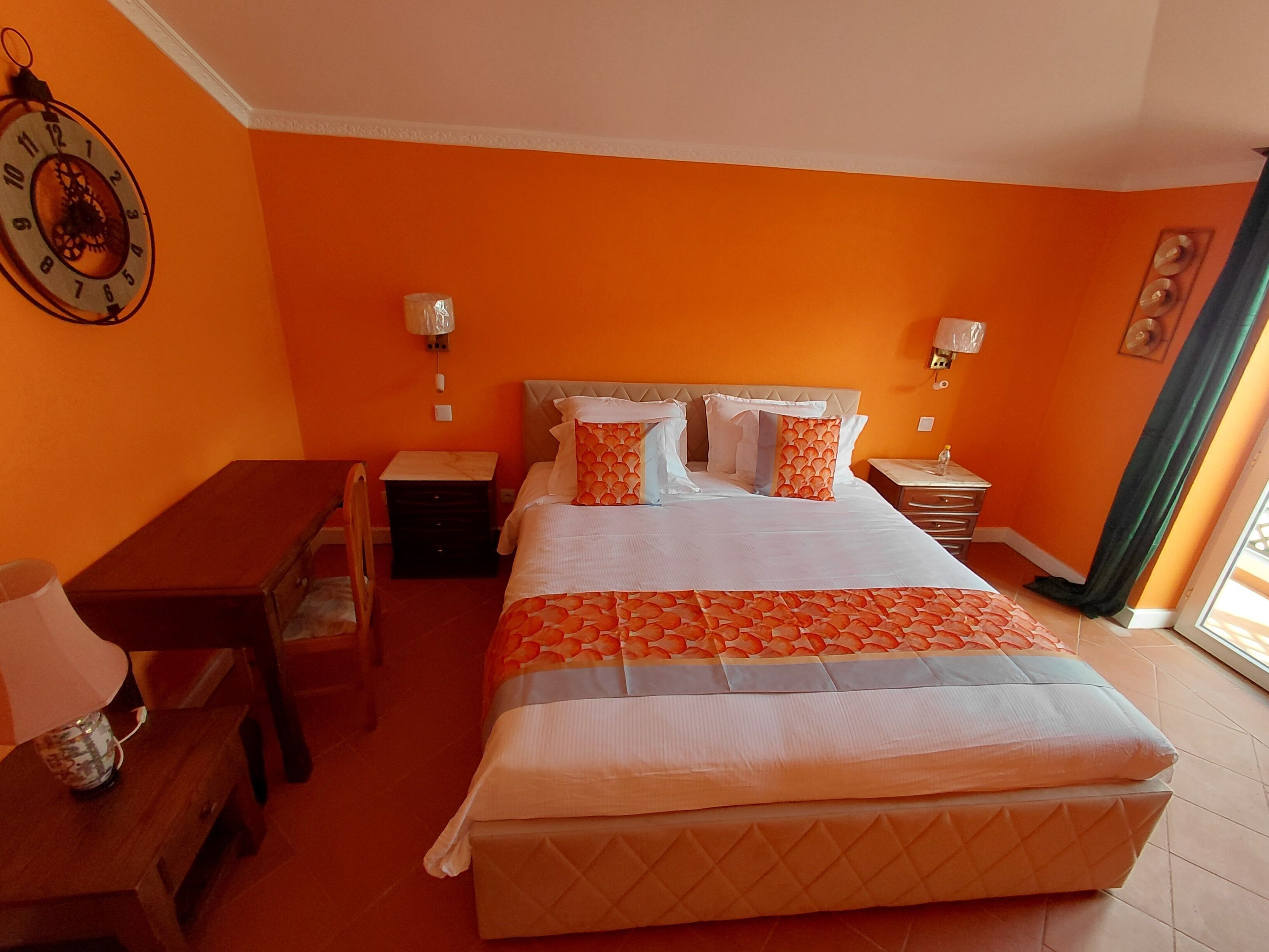 Quarto de hotel com parede laranja, cama com almofadas estampadas, mesa de cabeceira, cadeira e relógio na parede.