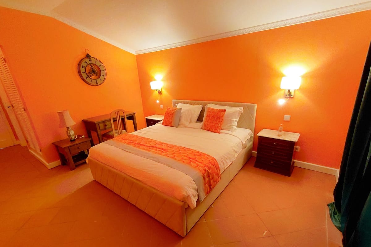 Une chambre à coucher avec un grand lit, deux tables de chevet, une petite table, une chaise et une horloge murale.