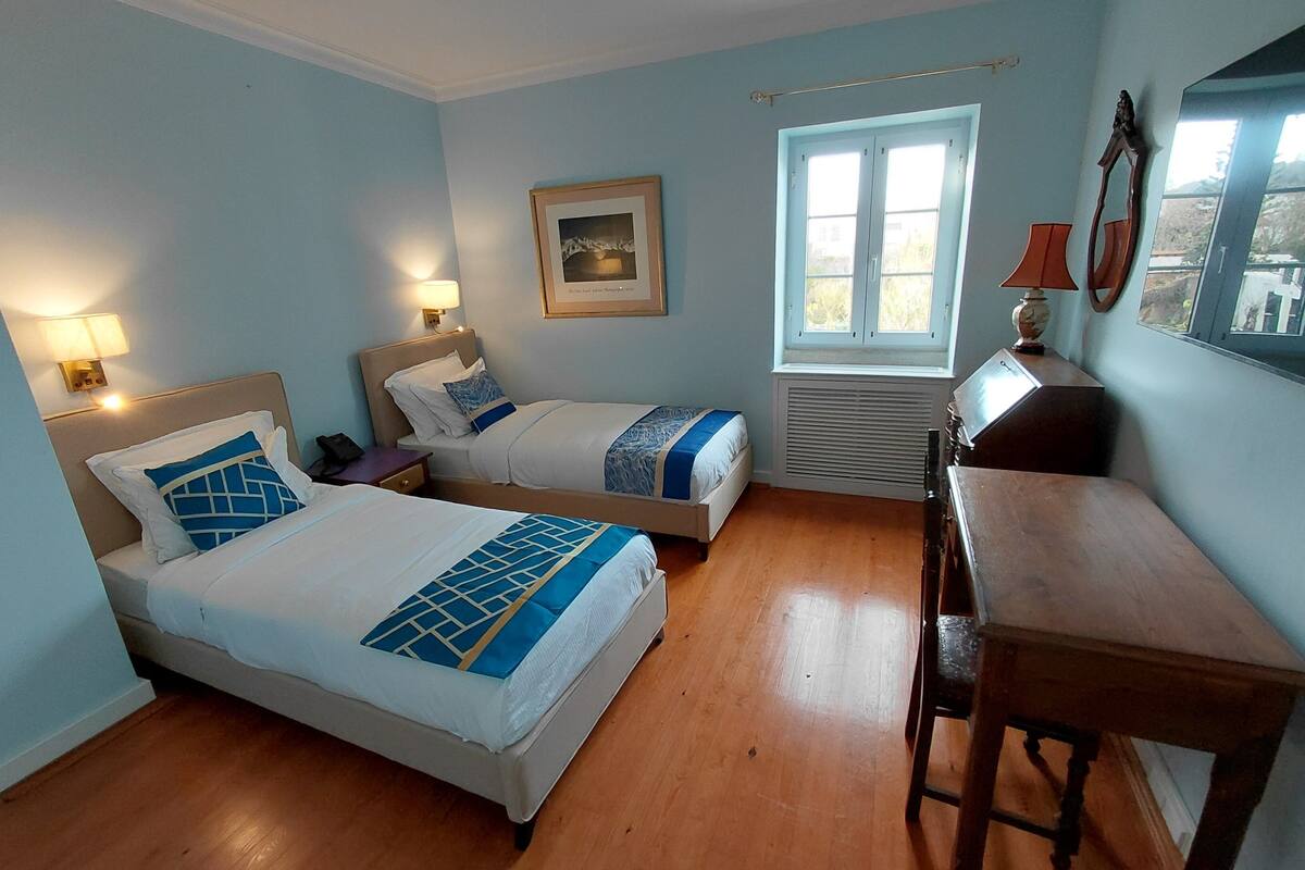 Une chambre d’hôtel avec deux lits, un bureau, une fenêtre et un tableau au mur.