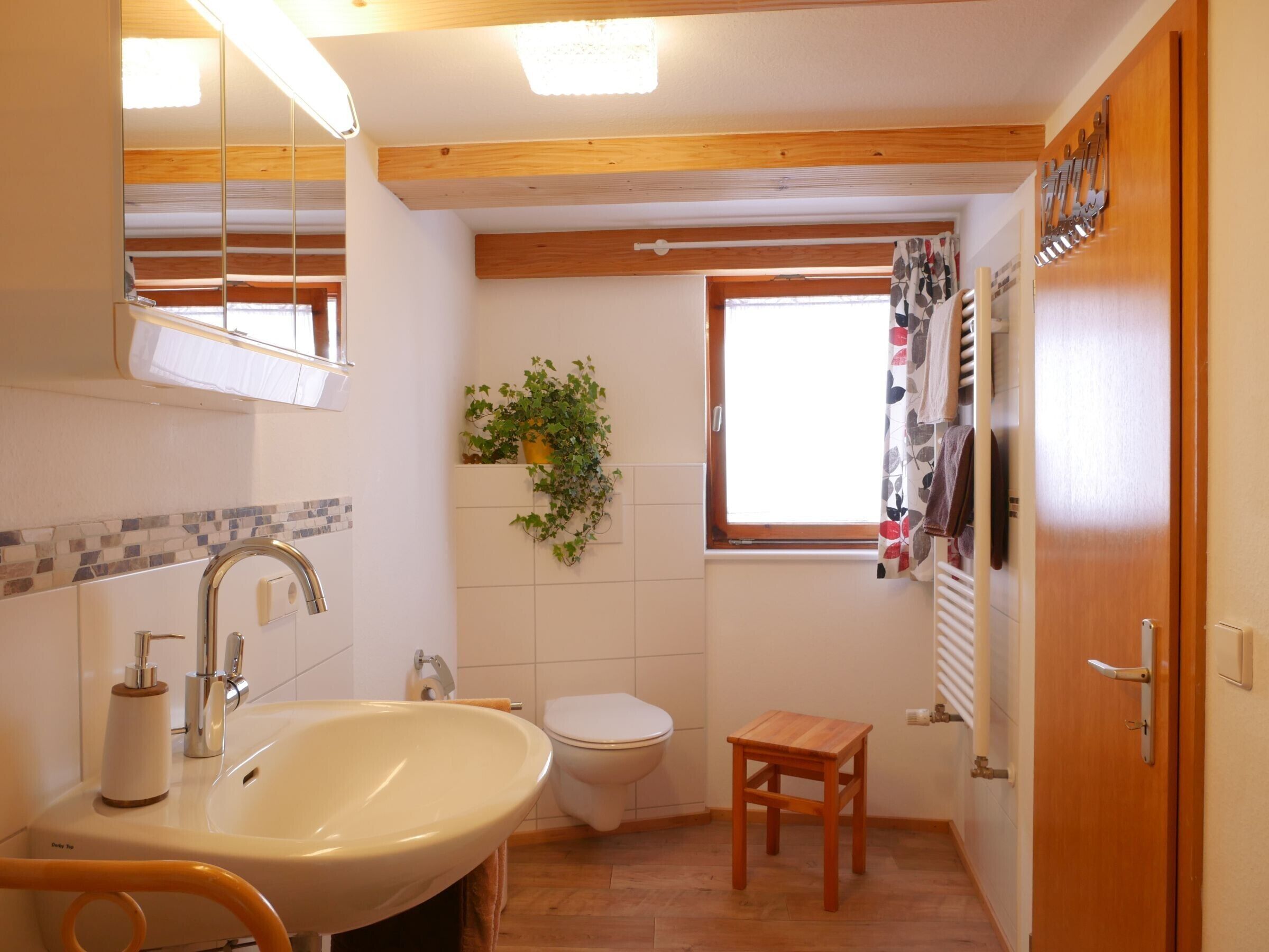 Un baño con lavamanos, inodoro, suelo de madera, un taburete pequeño de madera, una ventana con cortinas y un espejo.