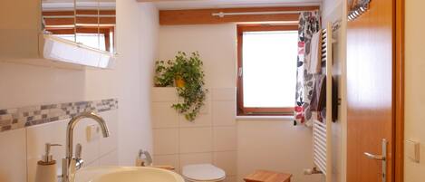 Un baño con lavamanos, inodoro, suelo de madera, un taburete pequeño de madera, una ventana con cortinas y un espejo.