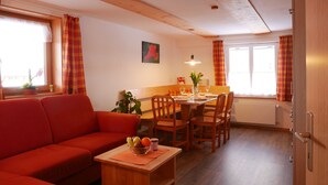 Ein gemütliches Wohnzimmer mit einem roten Sofa, einem Holzesstisch und Stühlen. Auf einem kleinen Tisch befinden sich eine Obstschale und eine Kerze.