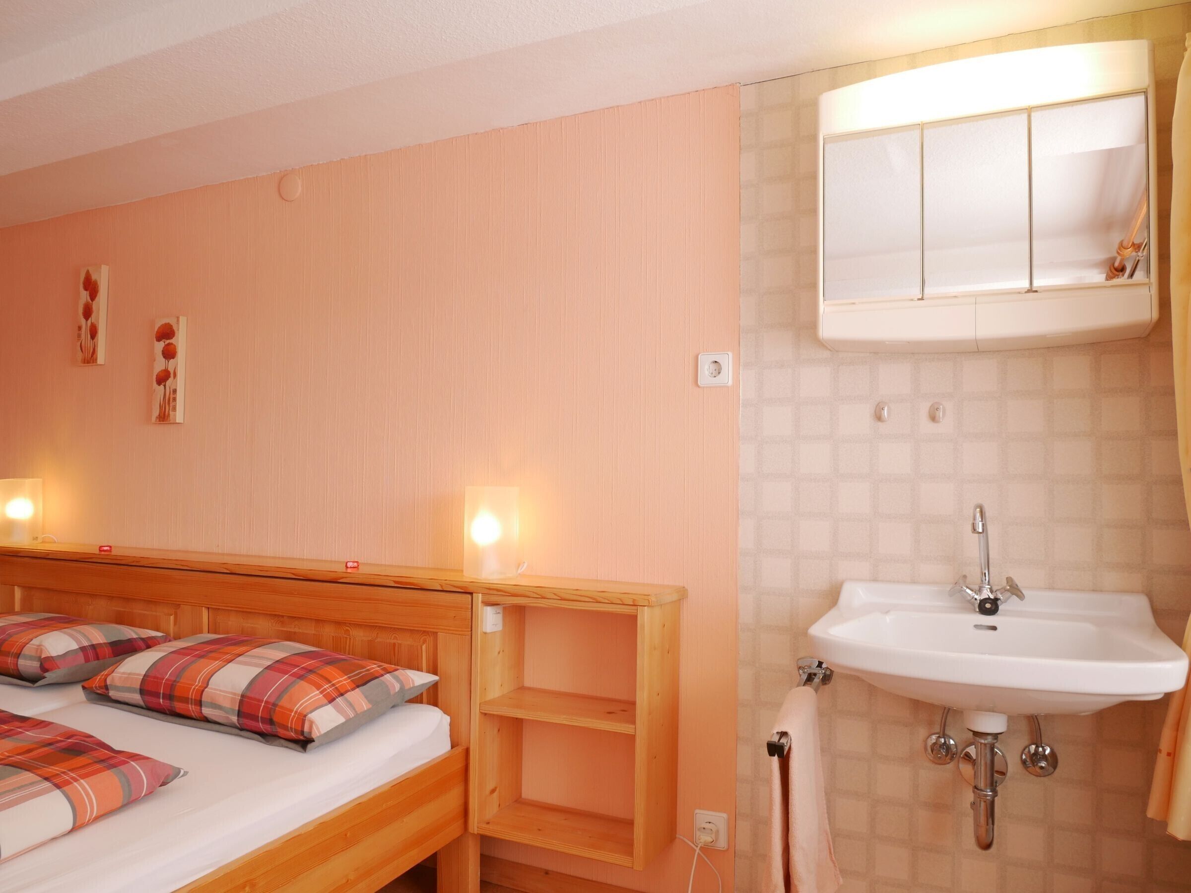 A bedroom with a wooden bed, a bedside table, a nightstand, a wall-mounted shelf, a bedside lamp, a wall-mounted mirror, and a bathroom with a sink and shower.