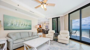 A spacious living room with a gray sofa, a white coffee table, and a beige armchair. A large painting of sailboats is on the wall, and the room has a large sliding glass door leading to a balcony with a view of the ocean. - Phoenix Ix 907 2 Bedroom Condo (Orange Beach)
