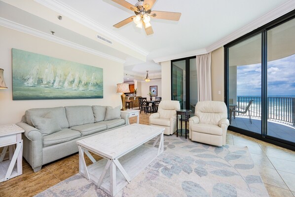 A spacious living room with a gray sofa, a white coffee table, and a beige armchair. A large painting of sailboats is on the wall, and the room has a large sliding glass door leading to a balcony with a view of the ocean. - Phoenix Ix 907 2 Bedroom Condo (Orange Beach)