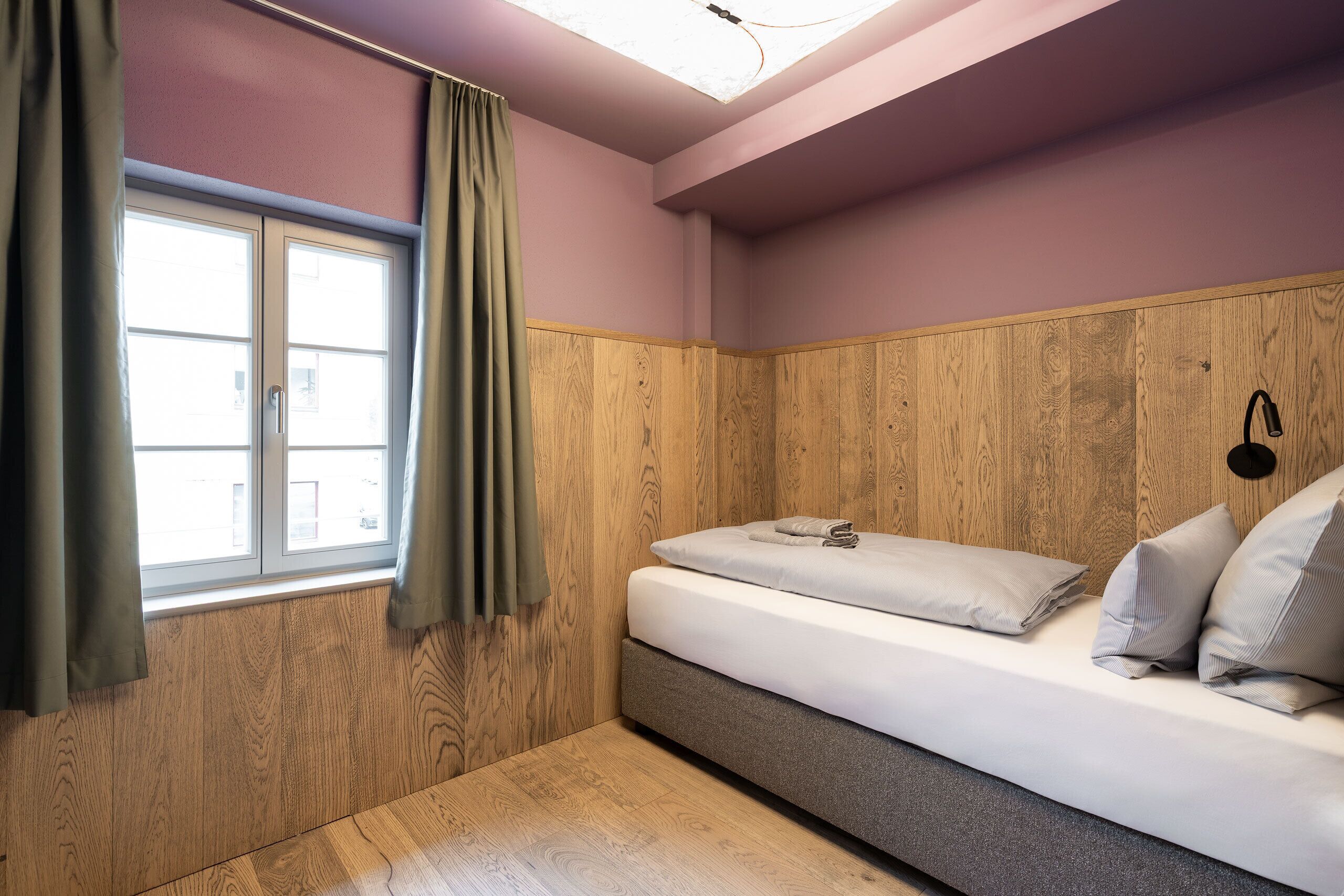 A small, cozy room with a bed, wooden paneling, a window with curtains, and a ceiling light.