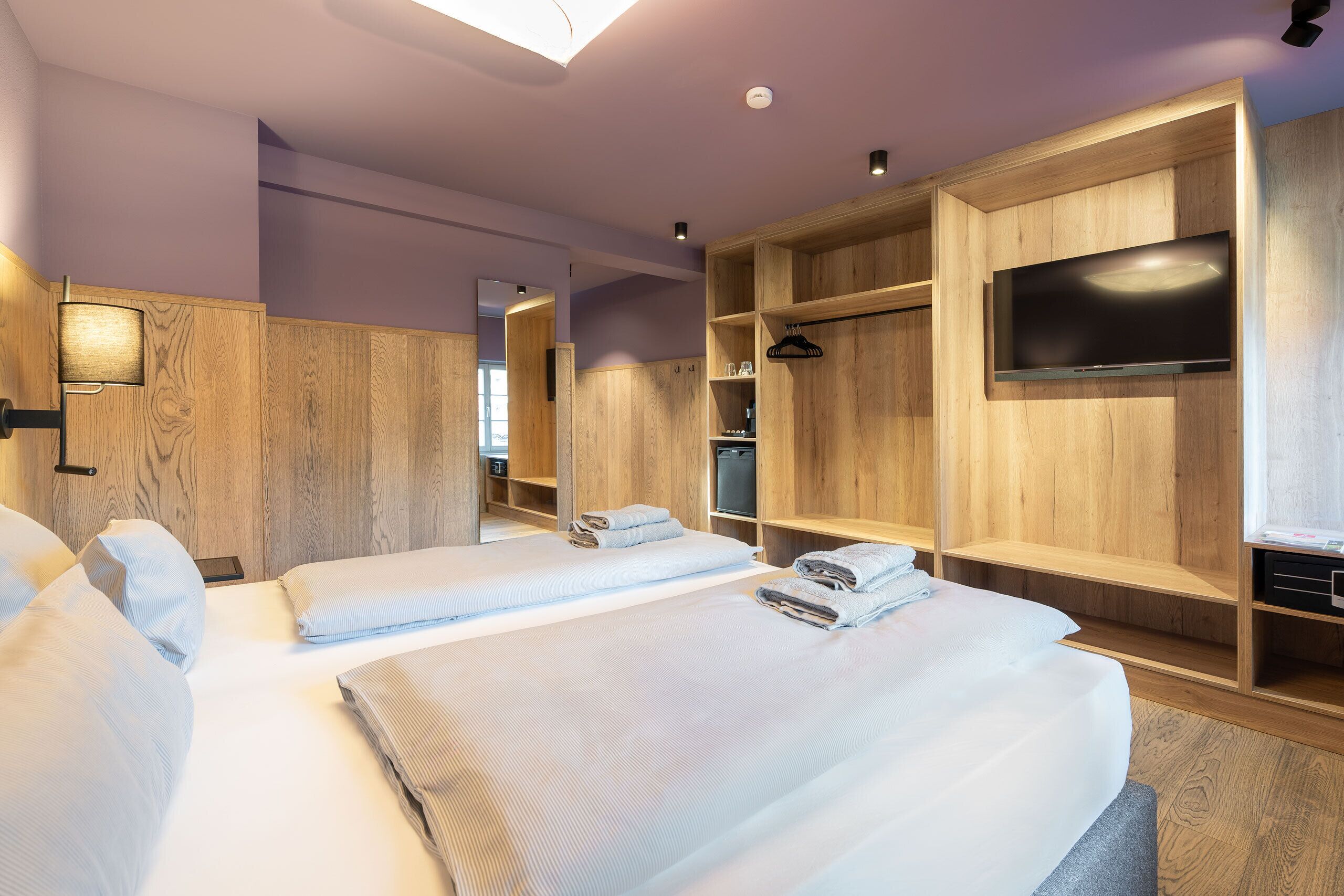Quarto de hotel com duas camas, uma televisão, estantes de madeira e um espelho.