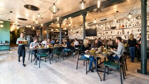 En restaurang med flera bord och stolar, en servitör som serverar mat och hyllor med diverse föremål i bakgrunden.