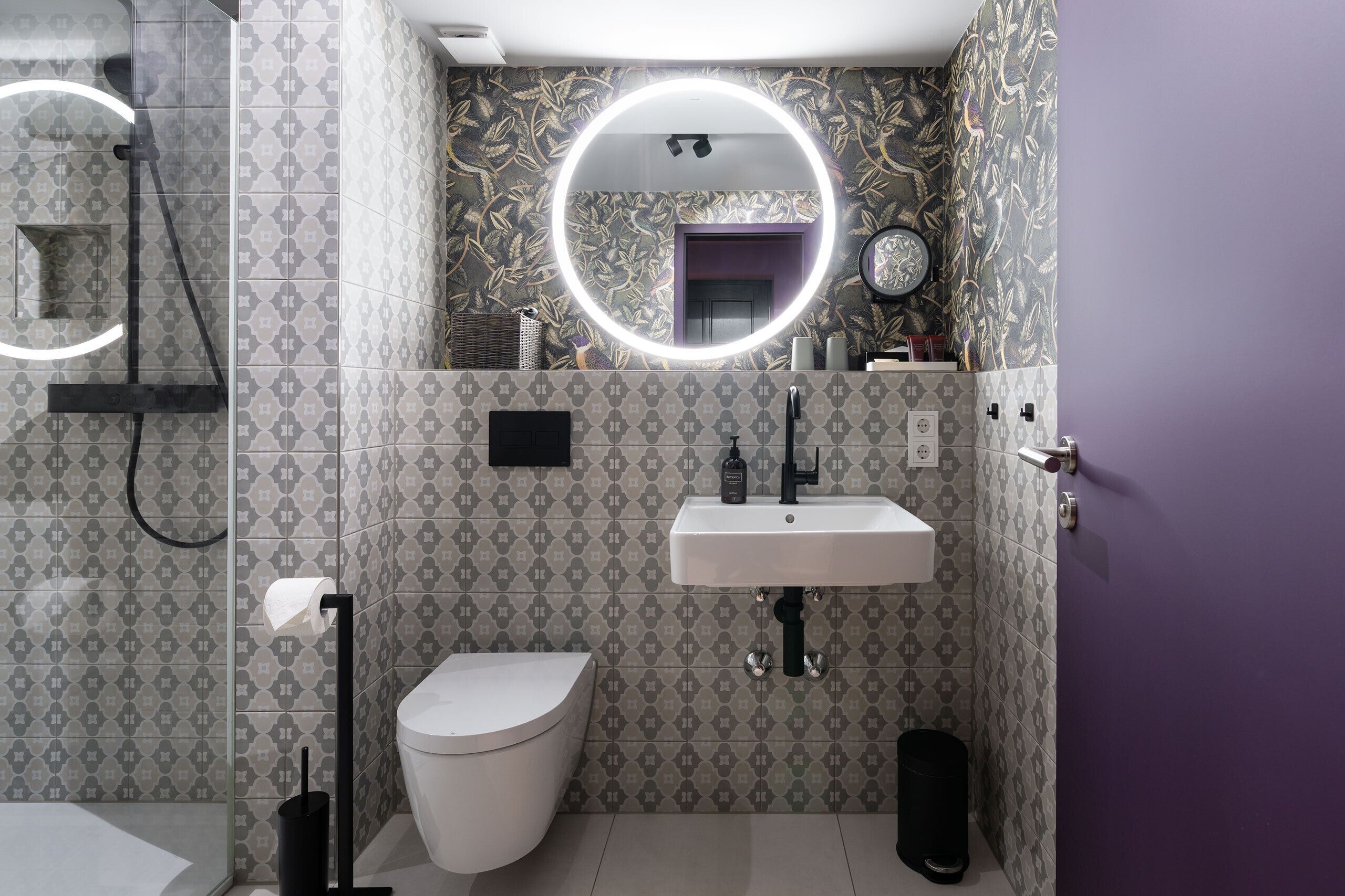 A bathroom with patterned wallpaper, a white toilet, a sink with a mirror, and a shower area.