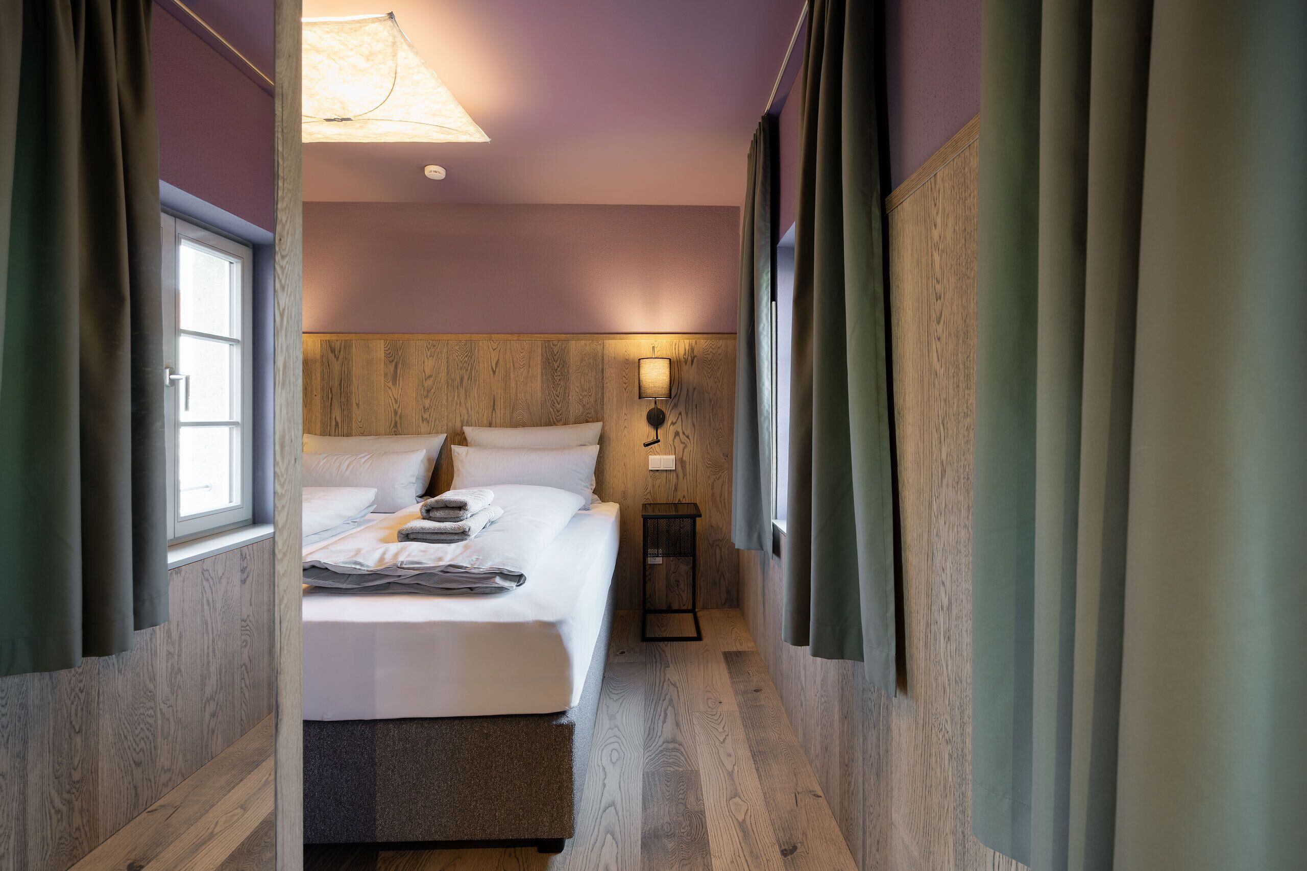 Habitación de hotel con cabecera de madera, cama con sábanas blancas, mesita de noche y lámpara de pared.