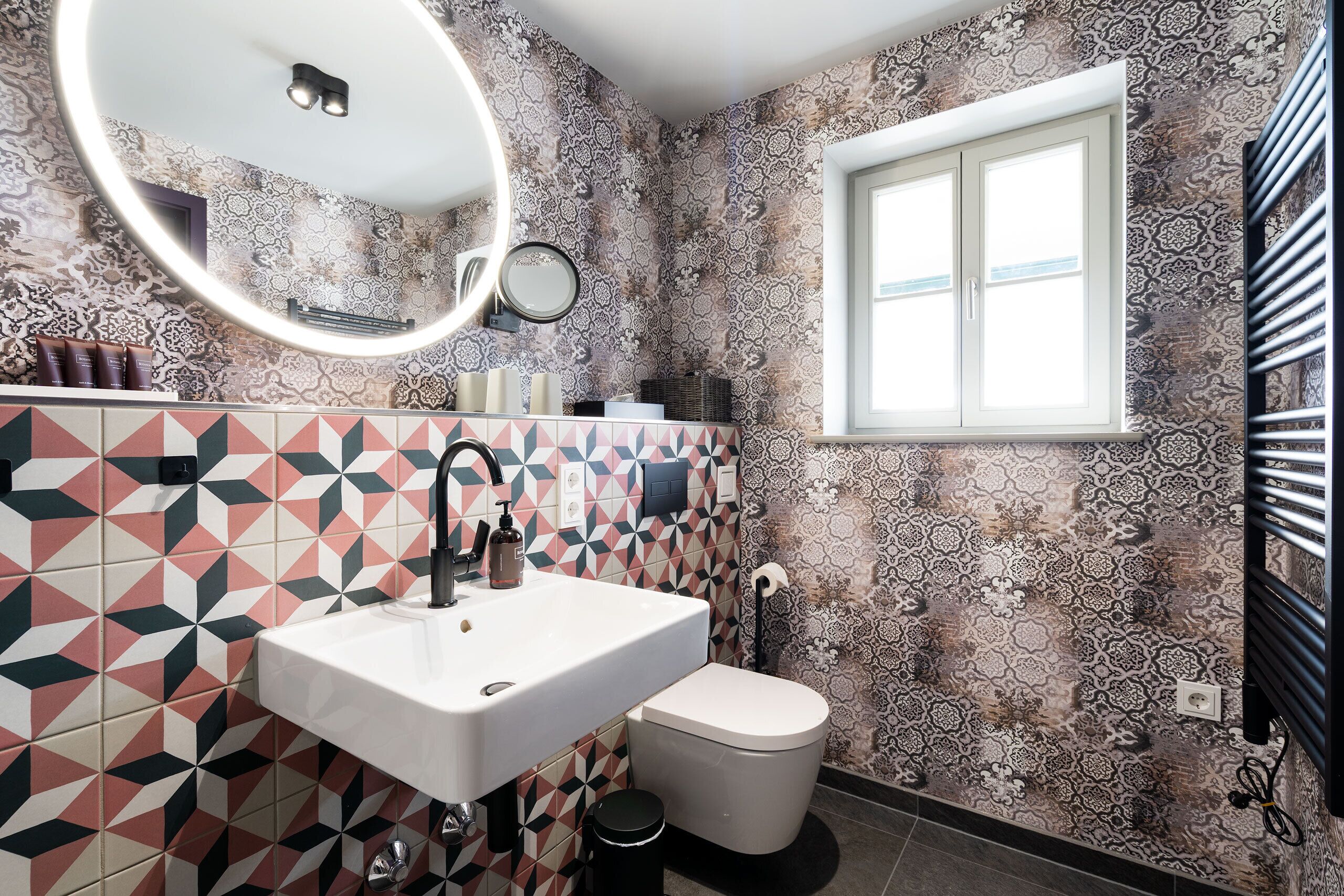 A bathroom with patterned tiles, a white sink, a toilet, a mirror, and a window.